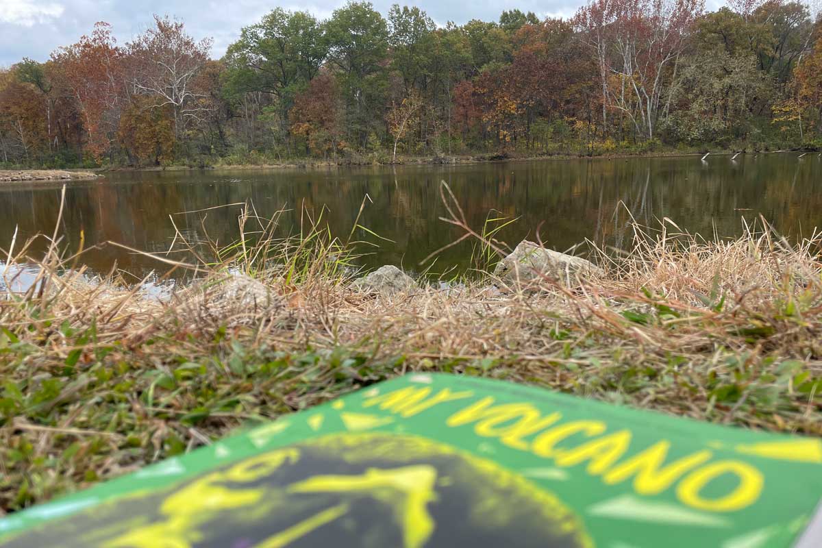 a book lies in the grass on a fall day. The book is blurry in the foreground and reads "My Volcano." A line of trees is in the background, and a rainy pond is in between.