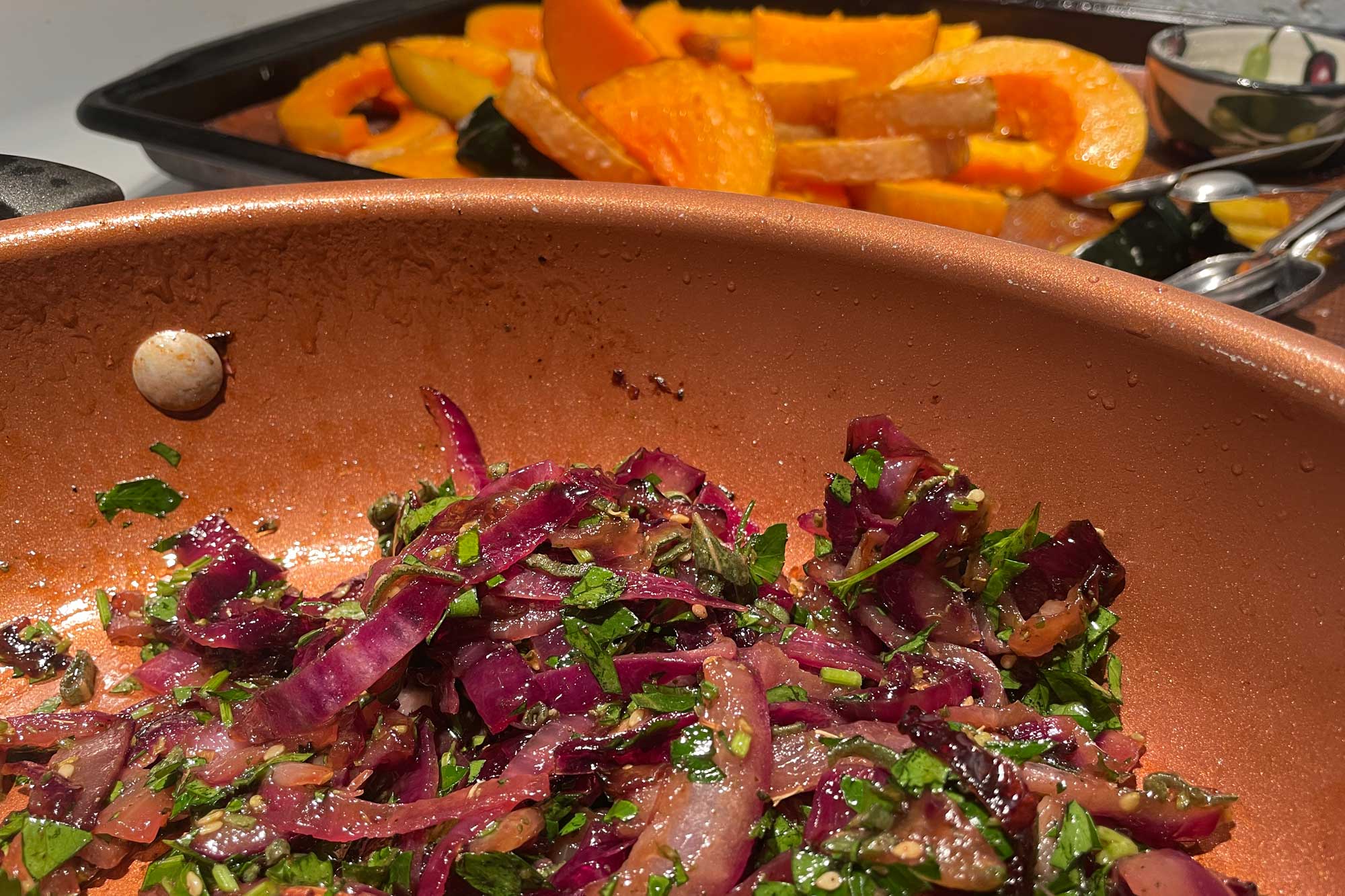 close-up on a pan filled with green herbs and red onions, with orange squash in the background. The vibe is fresh, active, and complete.